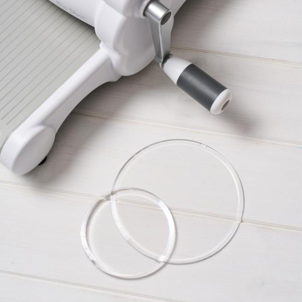 Clear Cutting Pads, Round, 2 Sizes ... by Sizzix. Clear circular plastic cutting plates to use with Sizzix die cutting templates and embossing folders. Sizes approx 5" x 5" (127mm in diameter) and 3 1/2" x 3 1/2" (88mm in diameter), each is 3mm thick (one of each size). This pack of Sizzix Cutting Pads includes a wonderful pair of round discs, circular cutting plates to use in your die cutting adventures. Photo of the Cutting Pads on a wooden table next to a Sizzix Big Shot machine (not included).