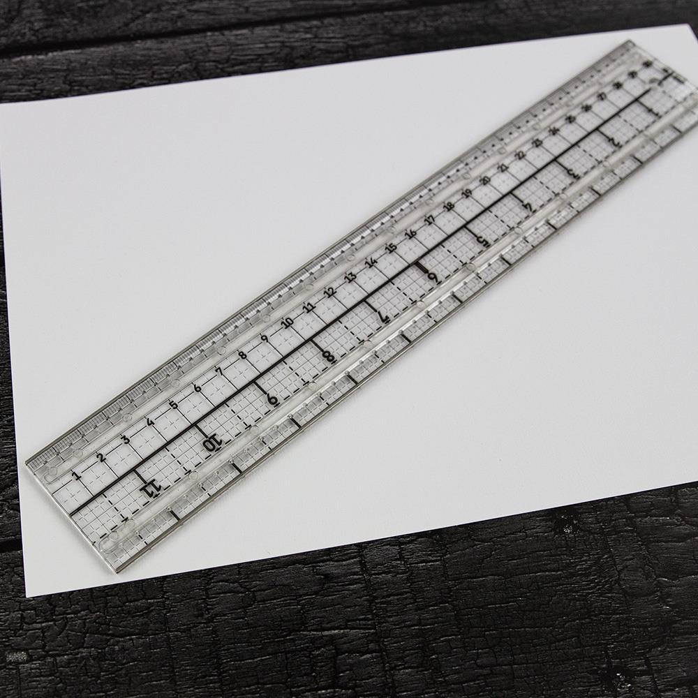 Media Ruler ... by Tim Holtz and Tonic Studios. Clear plastic ruler with precise measurements in both metric and imperial, dual steel cutting edges, non-slip backing, 2" wide and 12" long. Photo of the ruler out of the box, in the wild being used with a white sheet of paper.