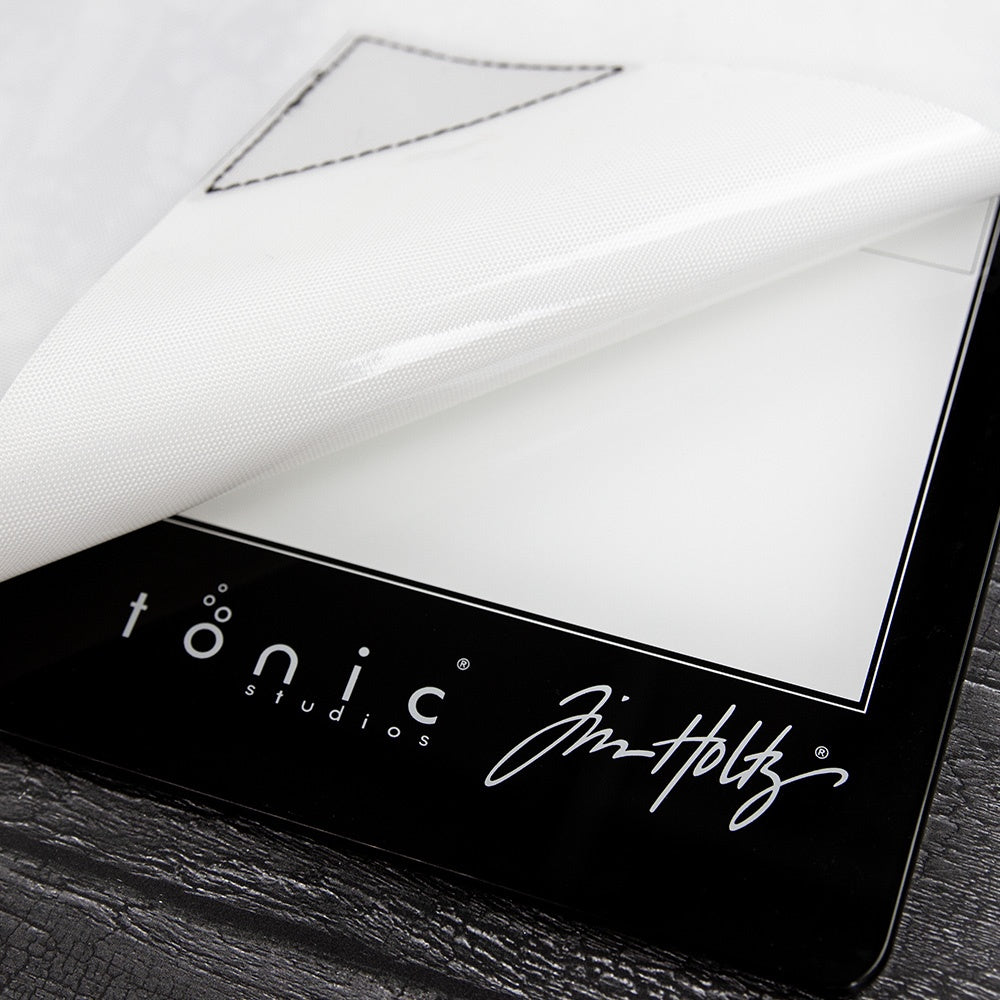 Small (Travel Sized) Media Surface Mat by Tim Holtz and Tonic Studios ... Travel Sized 15.5" x 10" heavy duty, non stick and heat resistant craft workspace with weighted reinforced corners ... designed to use as a stand alone craft mat or with Tim's Travel Sized Glass Media Mats (all sizes also available, each sold separately) as well as on its own. Photograph showing a closeup photo of the back of the mat on top of the glass mat showing the brand logos.