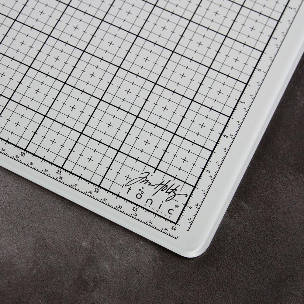 Closeup photograph of the lower right corner of the Bright White Glass Media Mat, Travel Sized - showing measurements, gridlins and the logos of Tim Holtz and Tonic Studios. This fantastic multi-functional tool at the portable size of 10.25" x 15.75", is ideal for creating and crafting in your workspace, whether in a studio, craft corner at home or during lunch at the office ... it is stylish and practical. It includes a storage case made of similar fabric to wetsuits, ensuring the glass is protected.