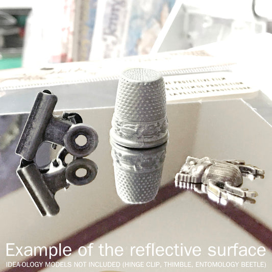 Mirrored Sheets ... Idea-Ology by Tim Holtz - adhesive backed sheets of highly reflective material that can be cut or used whole, for use as embellishments for scrapbooking, papercrafts, craft decor, display makes, creative visual arts and mixed media projects (TH93029). 2 (two) sheets, each 6" x 9" (15cm x 23cm). Use this mirrored surface to create miniature mirrors, silver trays, reflective element in your artwork. Photograph unedited, showing the mirrored sheet reflecting a metal clip, thimble and bug.