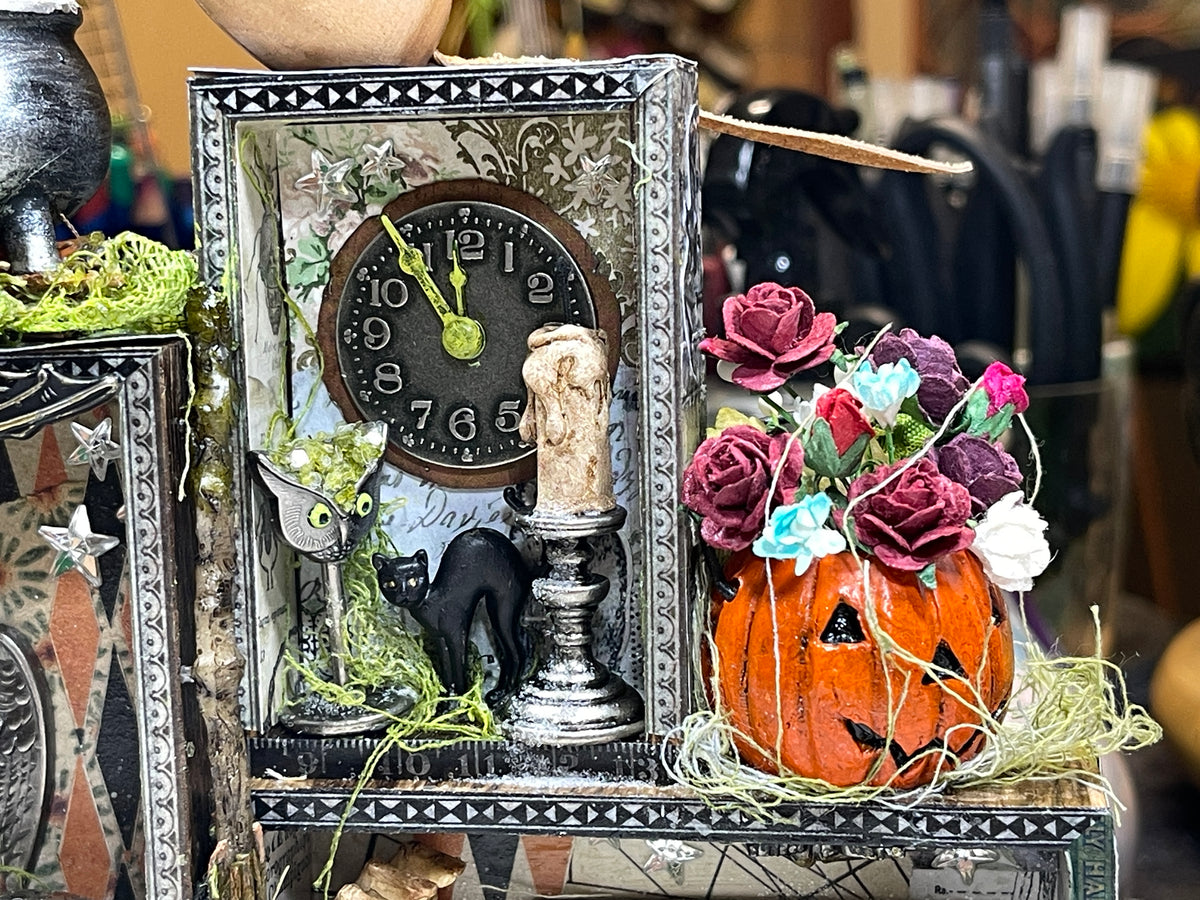 A decorative Halloween-themed tabletop display made by Jenny James using Tim Holtz Idea-ology. Features a vintage-style clock with an owl figurine, a drippy candle on candlestick, a small black cat, and a carved orange pumpkin with smiling face filled with paper roses in purple, pink, blue, and white.