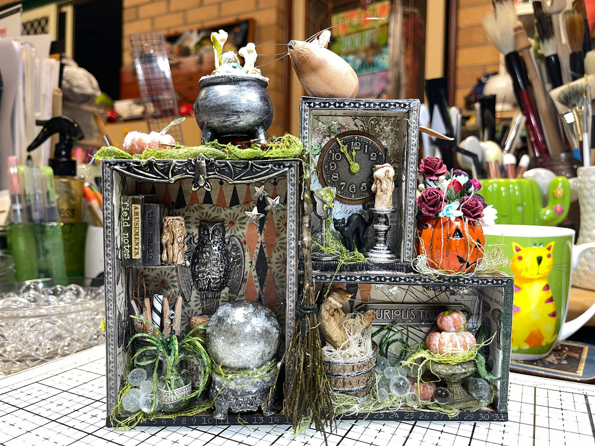 A decorative 3D collage mixed media made using 4 layered Tim Holtz Vignette Trays. Made by Jenny James, featuring gothic and whimsical elements, including a wooden mouse, cauldron of eyes and bones, miniature broom, drippy candle, shelf of tiny books, vintage clock, dried flowers, and moss, displayed on a craft table.