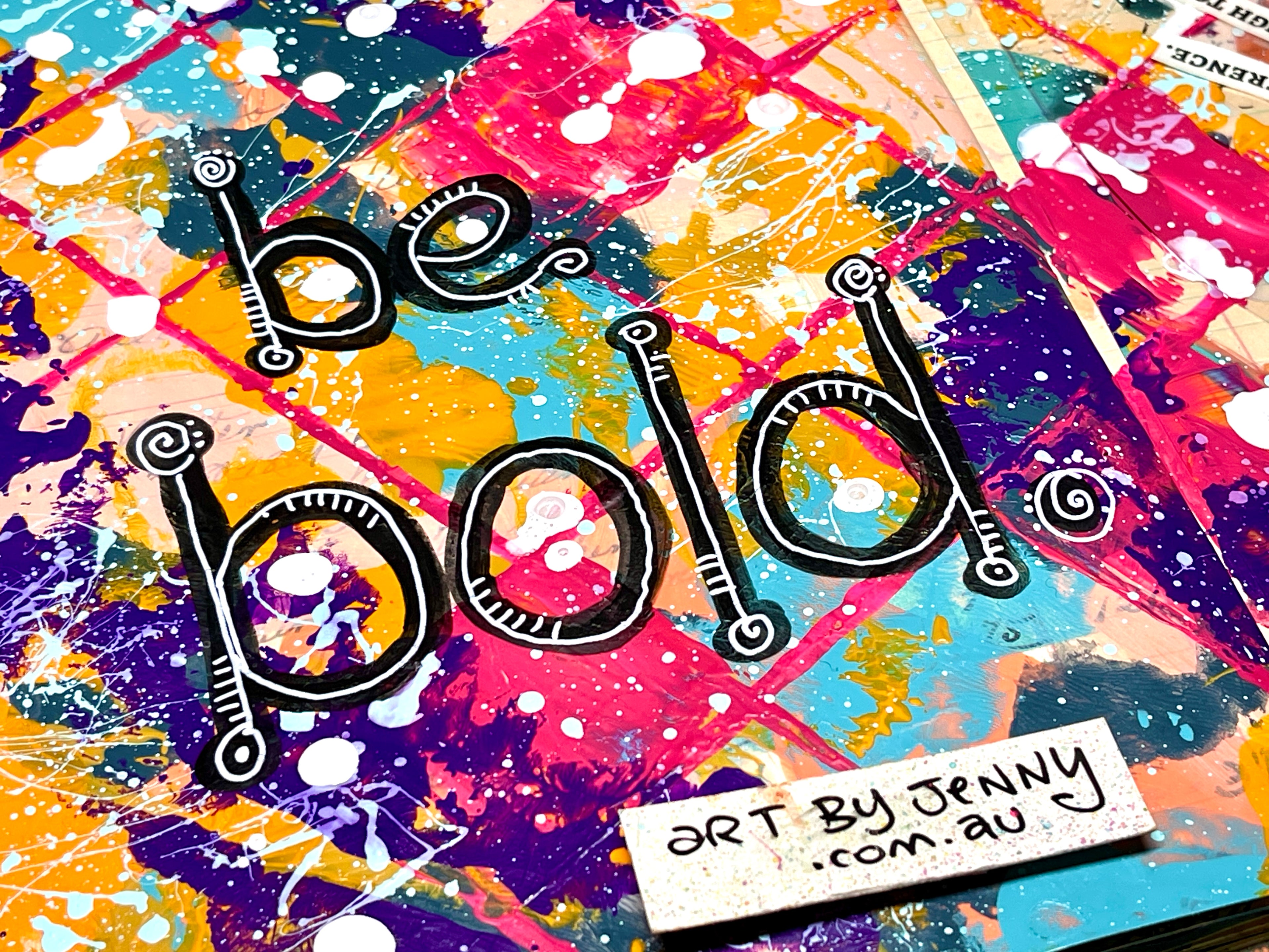 A colorful abstract art print featuring the hand-lettered text 'be bold.' in black outline over a vibrant paint-splattered background in pink, blue, yellow, orange, and purple.