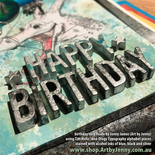 A close up detail of a birthday card made by Jenny James (Art by Jenny) featuring the Tim Holtz Idea-Ology Typography letters and stars. Stamped dragon in the background is Derpy by Brett Weldele.