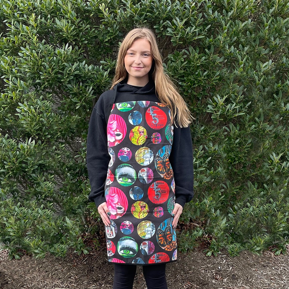 Young person wearing - Apron ... by Dyan Reaveley's Dylusions and Ranger. Designed with the same imagery as we have seen on the Dylusions bags, Dyalog cover and accessory cases - featuring various sized circles with bold beautiful colourful artwork over a black background. Adult sized, large, made of canvas, bound with black edging and an adjustable neck strap. An excellently sized and shaped coverall for those of us with larger shaped body parts! It also features two large pockets and long straps.