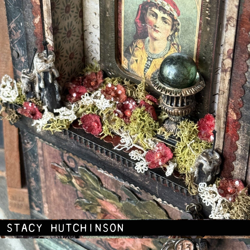 Detail of a beautiful Vignette frame by Stacy Hutchinson, showing a closeup view of the Gypsy, crystal ball, flowers and the blackened Tim Holtz Idea-Ology Drippy Candles. The photo is of a decorative handmade mixed media frame featuring a vintage-style portrait, a green crystal ball on an ornate stand, red paper Idea-Ology flowers, white lace, moss, Backdrops and Ephemera on the flat areas of the box.
