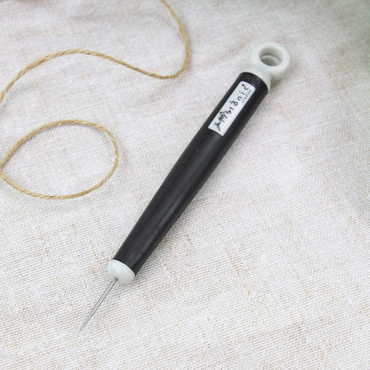 Retractable Craft Pick - by Tim Holtz and Tonic Studios ... this is a 5" long visual arts tool with a black Kushgrip rubber handle, hanging loop and easy to use switch. Use the long steel point for poking holes, lifting layers, clearing nozzles, extends up to 1.25 inches and holds safely in place at various depths. Photo from Tonic Studios, showing tool in use on a canvas background with coil of string.