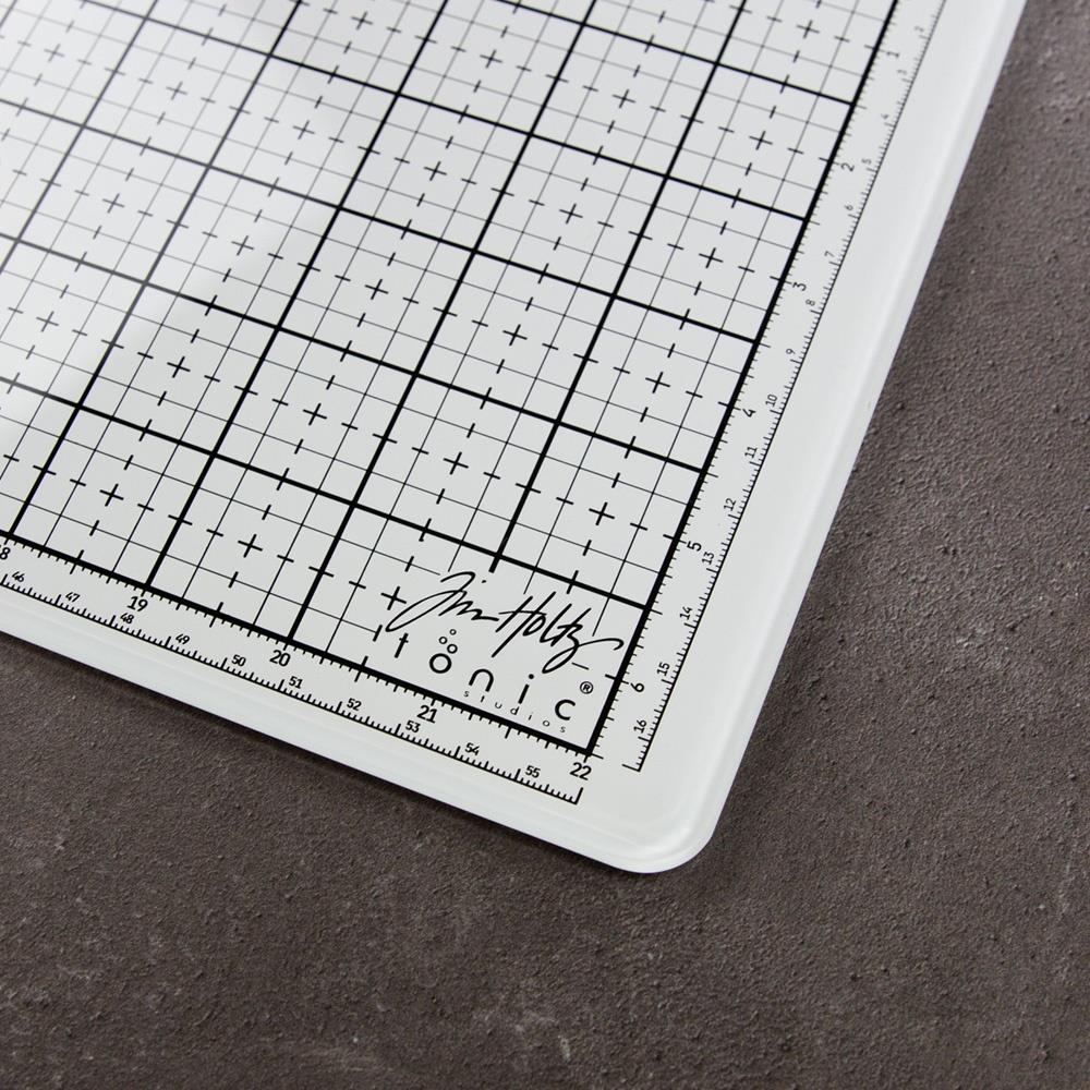 White Studio Glass Media Mat - Large Creative Surface 14"x23" ... by Tim Holtz and Tonic Studios. A bright white tempered glass mat with gridlines, silicon feet with both metric and imperial measurements marked around all the edge. Photograph of a closeup, lower right corner of the glass mat.