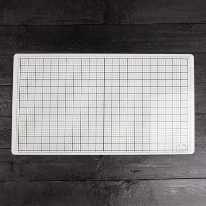 White Studio Glass Media Mat - Large Creative Surface 14"x23" ... by Tim Holtz and Tonic Studios. A bright white tempered glass mat with gridlines, silicon feet with both metric and imperial measurements marked around all the edge. Photograph showing the top surface of the glass mat on a timber tabletop background.